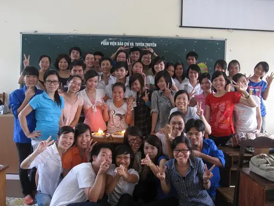 Nhóm đông người tụ quanh bàn có bánh và nến sáng trong lớp học, cùng nhìn máy ảnh và tạo dáng.