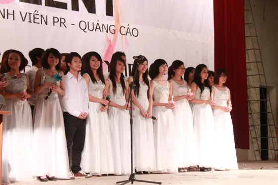 Nhóm người đứng thành hàng trên sân khấu cạnh chân micro, phần lớn mặc váy trắng dài.