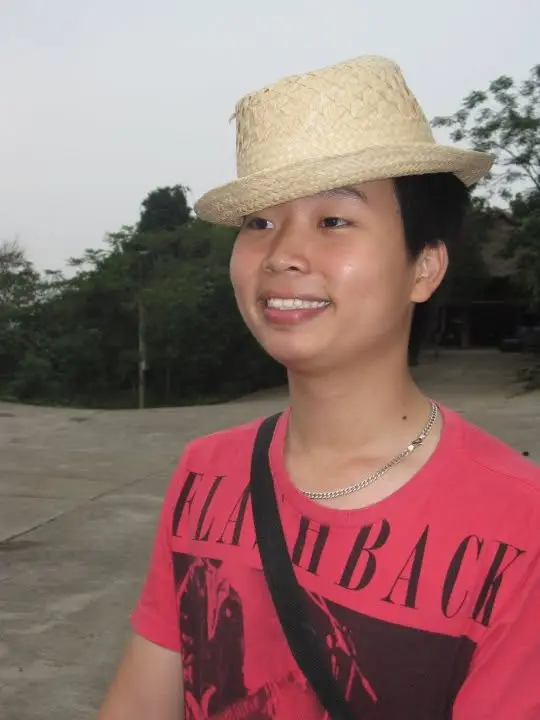 Một người đội mũ rơm, mặc áo phông đỏ và đeo dây túi chéo màu đen, mỉm cười trên sân ngoài trời.