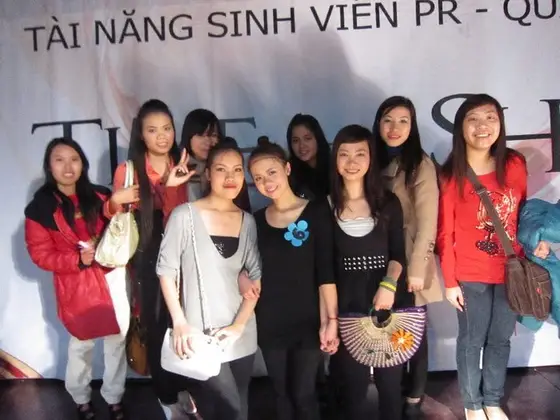 Nhóm người mỉm cười trước phông tài năng sinh viên PR, người phía trước mặc áo đen có hoa xanh cài ngực.