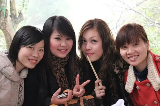 Bốn người ngồi sát nhau ngoài trời và mỉm cười, một người cầm điện thoại giơ tay chữ V, một người cầm đũa.