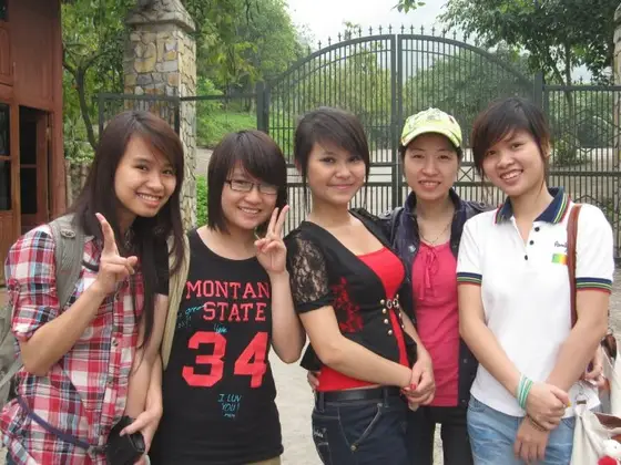 Năm người đứng sát nhau trước cổng sắt và cây xanh, hai người giơ tay chữ V, một người đội mũ xanh nhạt.